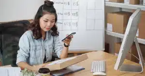 Imagem de uma mulher em seu home office com computador, celular, quadro branco e papéis em caixas aprendendo como ser mais organizada