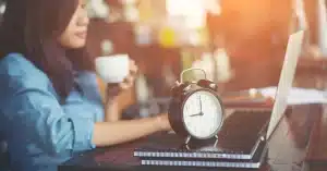 Imagem de uma mulher em frente ao seu laptop com uma xícara de café, cadernos e um relógio em cima da mesa otimizando o tempo