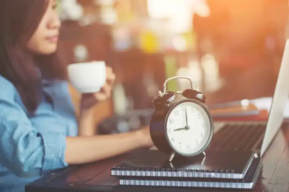 Imagem de uma mulher em frente ao seu laptop com uma xícara de café, cadernos e um relógio em cima da mesa otimizando o tempo