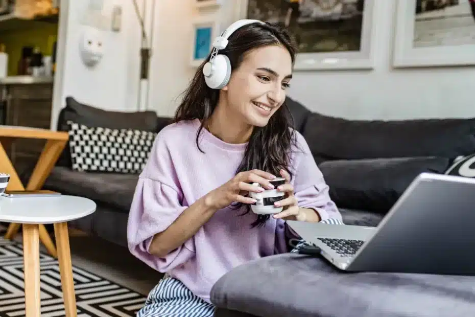 Mulher com fones de ouvido e segurando uma caneca, sentada no chão da sala com seu laptop, sorrindo para a tela com os resultados de ter aprendido como integrar Blog, Pinterest e YouTube